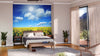 A bright bedroom features a bed facing the Field of Sunflowers Wall Mural, flanked by nightstands and potted plants. Sheer curtains cover balcony doors, letting light flood the wooden floor and cozy, neutral-toned decor.