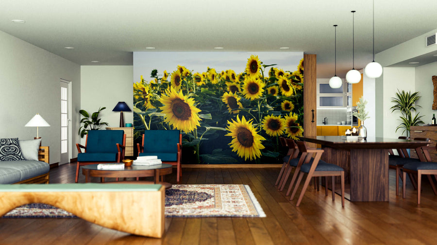 A modern open-concept living and dining room with wooden floors and mid-century furniture features the vibrant Field of Yellow Wall Mural as a focal point, adding color to the neutral space with green plants, pendant lights, and eclectic decor.