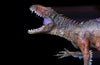 Close-up of a dinosaur figurine against a black background, with textured brown and green scales and an open mouth revealing sharp teeth and a purple tongue. Its red eyes and spiky ridges evoke the Fierce Prehistoric Predator Wall Mural