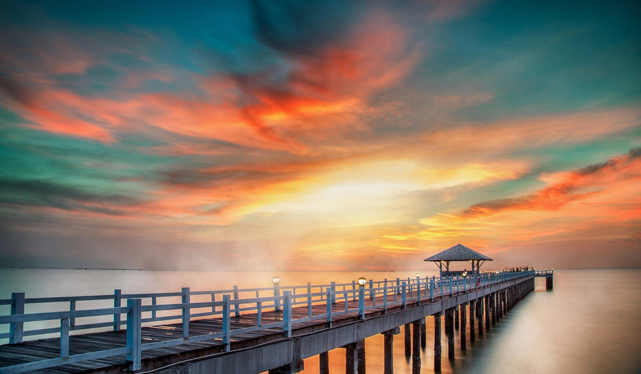 Modern open-concept living and dining room with wood floors. The Fiery Coastal Dusk Wall Mural of a sunset over a pier creates a striking focal point, while teal chairs, gray sofa, wooden table, and pendant lights add cozy contemporary charm.