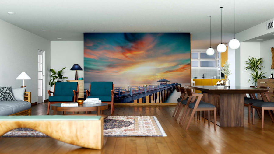 Modern open-concept living and dining room with wood floors. The Fiery Coastal Dusk Wall Mural of a sunset over a pier creates a striking focal point, while teal chairs, gray sofa, wooden table, and pendant lights add cozy contemporary charm.