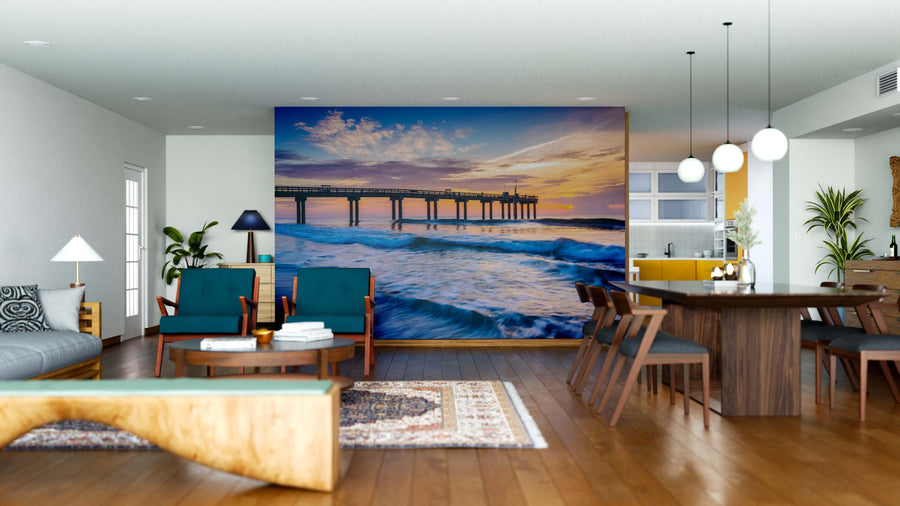 A modern open-plan living and dining room with wood floors and mid-century furniture features a large Fishing Pier Wall Mural. Ocean waves and colorful sunset clouds create a serene vibe, with the kitchen visible in the background.