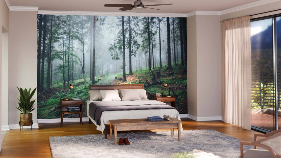 A cozy bedroom features the Foggy Forest Dirt Road Wall Mural behind a double bed with white bedding and a dark throw. Wooden nightstands, a plant, bench, gray rug, and floor-to-ceiling windows with sheer curtains complete the look.