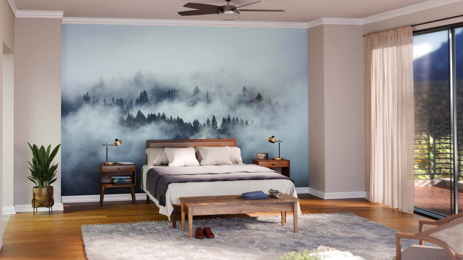 A modern bedroom features the Foggy Forest Through the Fog Wall Mural behind a wooden bed, nightstands with lamps, a bench at the foot, a gray rug, a plant, and sliding glass doors with sheer curtains welcoming sunlight.