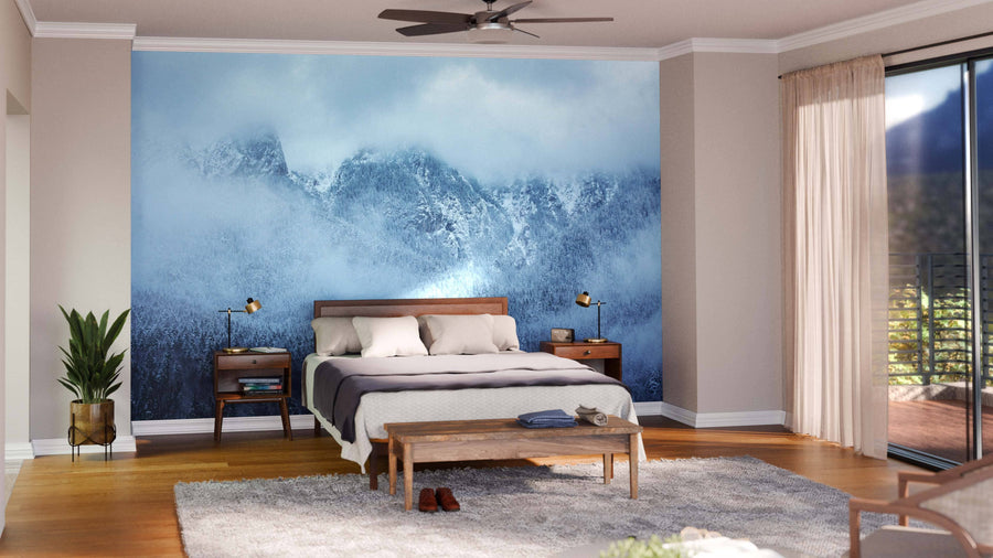 A modern bedroom features the Foggy Mountain Peaks Wall Mural behind the bed, wooden furniture, a soft area rug, a potted plant, and large windows with sheer curtains that allow natural light to fill the space.