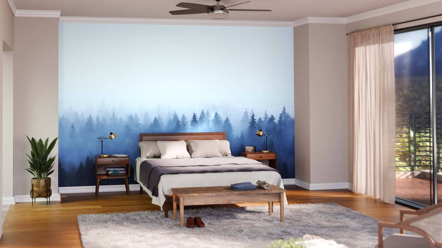 A bedroom with a bed, two nightstands, and lamps features the Foggy Pine Forest Wall Mural behind the bed. A wooden bench sits at the foot, with a rug and potted plant to the left, plus sliding glass doors with curtains on the right.