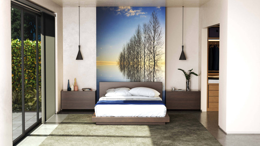A modern bedroom features a low wooden bed with white bedding, dark nightstands, and pendant lights above. Behind the bed, the Forces of Nature Wall Mural depicting leafless trees by a lake at sunset adds drama. Sliding glass doors open to the left.