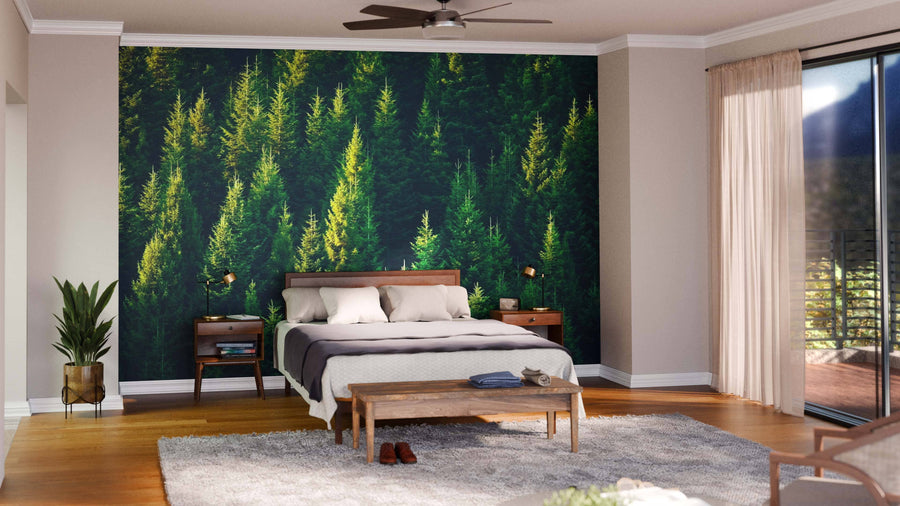 A cozy bedroom showcases the Forest Tree Tops Wall Mural behind a wooden bed, with a ceiling fan, nightstands and lamps, bench, plant, gray rug on hardwood floors, and sliding glass doors leading to the balcony.