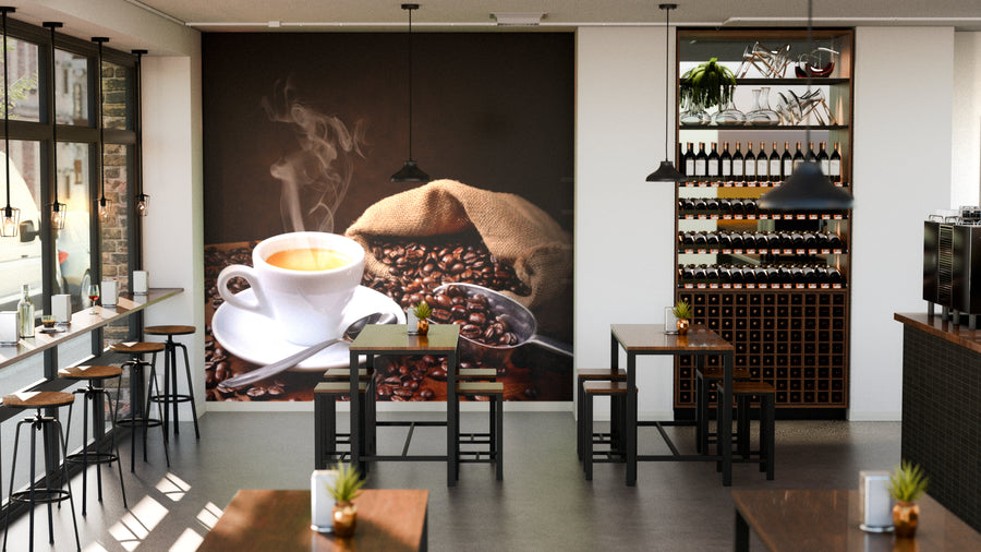 A modern coffee shop with wooden tables, chairs, and potted plants features the Fresh Roasted Coffee Beans Wall Mural showing a steaming cup of coffee. Bottles line shelves, while sunlight pours in through large windows on the left.