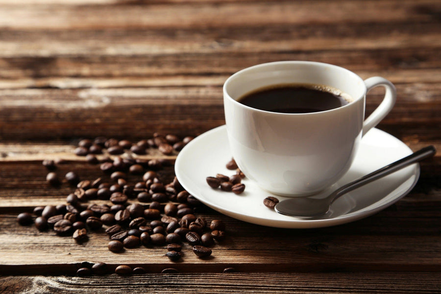 A modern cafe interior with wooden tables, black chairs, potted plants, and large windows features the Freshly Brewed Black Coffee Wall Mural—a close-up of a white cup of coffee with beans on rustic wood—plus pendant lights overhead.