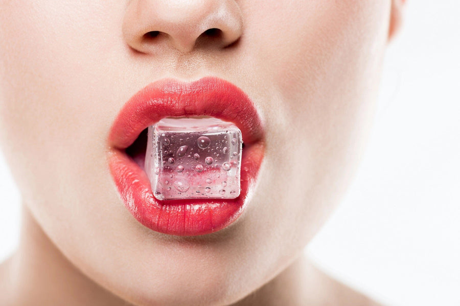 A close-up of a person with red lipstick holding a clear ice cube between their lips, set against the Frosted Lips Wall Mural. The smooth skin and water droplets on the cube stand out against the plain white background, enhancing the vivid lip color.