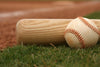 A close-up of a wooden baseball bat and a white ball with red stitching on green grass, set against a blurred stadium infield—Game Day Ready Wall Mural, ideal for adding sporty flair to your space with easy peel-and-stick application.