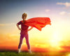 A child confidently stands on grass facing a vibrant sunset, akin to art on "Girl Plays Superhero" wall mural. Wearing a red cape flowing in the breeze and hands on hips, the orange and blue sky enhances the heroic mood, symbolizing strength and imagination.