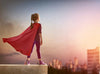 A young girl stands on a ledge, her red cape fluttering against a sunset city skyline. In purple leggings and sparkly sneakers, she captures the spirit of adventure—an image as stunning as the "Girl Superhero Wall Mural"