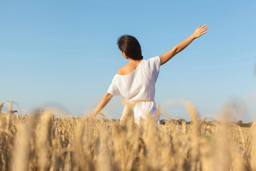 A modern open-concept living and dining room with wood floors and mid-century furniture features the Girl in Wheat Field Wall Mural, showing a woman in a white dress standing in wheat under a clear blue sky, one arm raised.