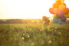 A young girl in a yellow dress joyfully runs through a sunlit grassy field with balloons, embodying the serenity of Happy Girls Wall Mural, where warm sunlight and distant trees under a clear sky enhance the scene.