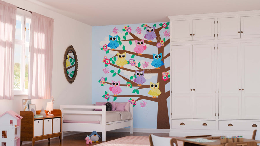 A children’s bedroom features the Owls on a Tree Wall Mural, showcasing cartoon owls and pink flowers. The cozy space includes a white bed with pink bedding, wardrobe, small table, mirror, dollhouse, and floor toys.