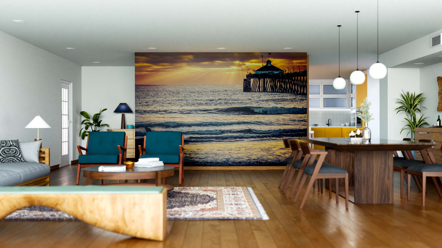 Modern open-plan living and dining area with wooden floors, teal and wood furniture, pendant lights, and the Golden Coast Horizon Wall Mural depicting a sunset over the ocean with a pier. Area rugs and decor add cozy style.