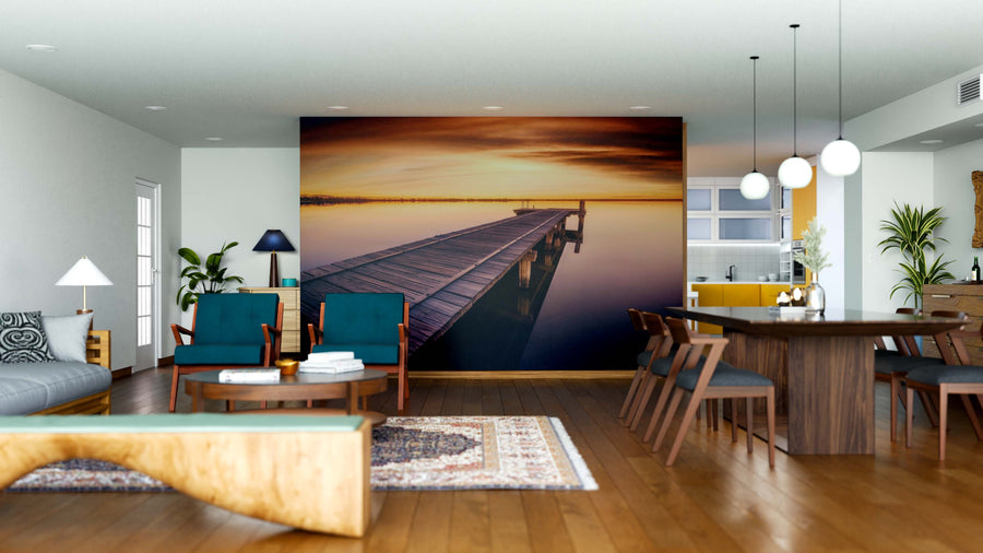 A modern living and dining area features wooden floors, blue and wood furniture, pendant lights, plants, and the Golden Dock Wall Mural—a peel-and-stick design of a pier at sunset—creating a serene focal point in the open space.