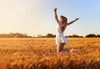 A woman in a white dress kneels joyfully with arms raised in a wheat field under the clear blue sky, reminiscent of the "Golden Field Bliss Wall Mural." Her cascading hair is highlighted by the warm sun against vibrant wheat and serene trees in the background.