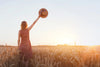 A woman in a sleeveless floral dress stands in a golden wheat field at sunset, raising a ribboned straw hat skyward as she faces away, embodying the tranquility captured in our Golden Fields Wall Mural to bring serene beauty into any space.