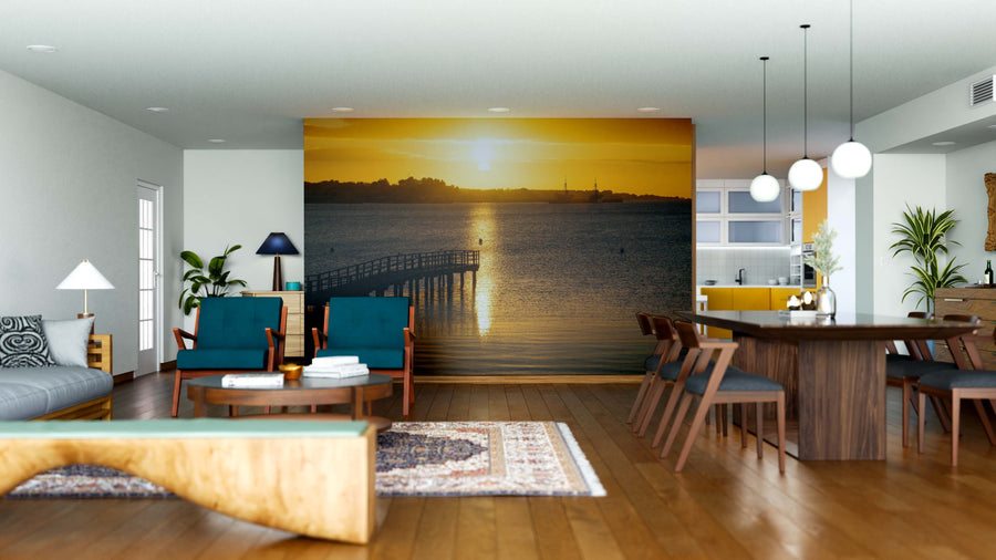 A modern, open living-dining room with wooden floors and mid-century furniture features the Golden Horizon Wall Mural, showcasing a sunset over water with a pier. Pendant lights above the table and greenery complete the inviting space.