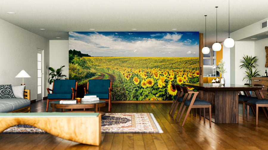A modern living and dining room with wooden floors, blue and wood furniture, and the Golden Horizon Trail Wall Mural, featuring a bright sunflower field under a blue sky. Pendant lights, houseplants, and rugs create a cozy, inviting space.