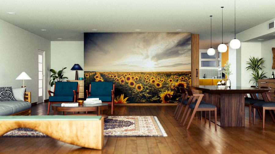 Modern open-plan living and dining room with wooden floors and furniture features the Golden Hour Bloom Wall Mural, showcasing a sunflower field at sunset. Blue armchairs, rugs, plants, and pendant lights add to the cozy, artful vibe.