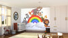 A bright nursery features the Happy Animal Rainbow Wall Mural with cartoon jungle animals—elephant, giraffe, zebra, and lion under a rainbow. The space includes a crib, toys, baskets on the wooden floor, and sunlight streaming through curtains.