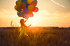 A young girl in a yellow dress holds colorful balloons in a field at sunset, with the sky's orange and pink hues resembling the "Happy Girl Wall Mural" The vibrant balloons beautifully contrast against the serene backdrop.