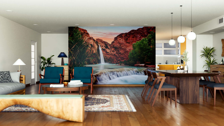 A modern open-plan living and dining area features wooden floors, teal armchairs, a wooden dining table, and the striking Havasu Falls Wall Mural depicting scenic waterfalls and red cliffs, accented by plants, a rug, and pendant lights.