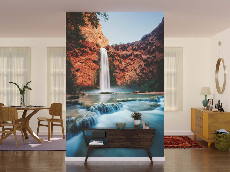 A living room with wooden floors, mid-century furniture, and a large Havasu Falls Arizona Wall Mural displays a vivid waterfall flowing into a turquoise pool amid red cliffs, bringing nature and tranquility to the modern space.