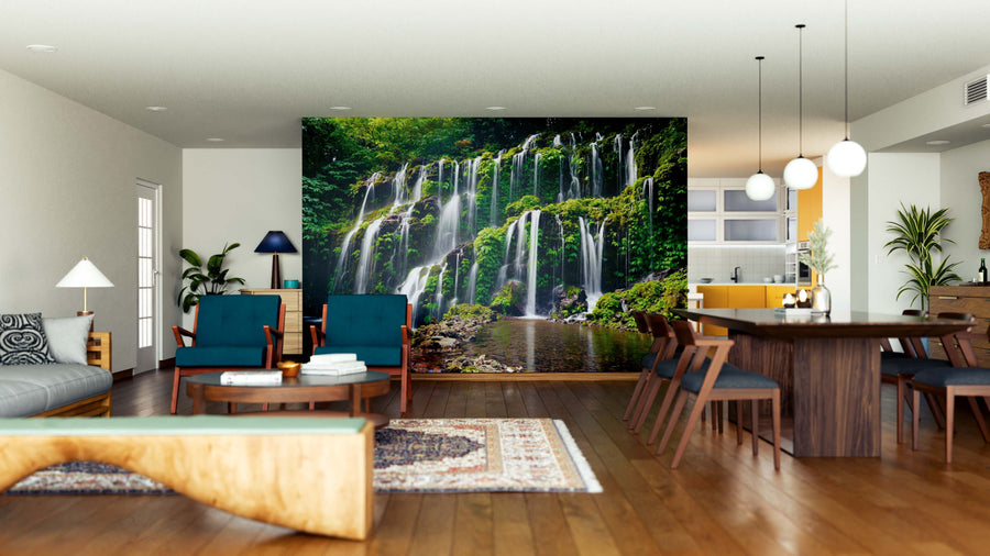 A modern open-plan living and dining room with wood flooring, teal chairs, a tan sofa, and the striking Hidden Waterfalls Wall Mural. The dining area features a wooden table, pendant lights, and a kitchen with yellow cabinets in the background.