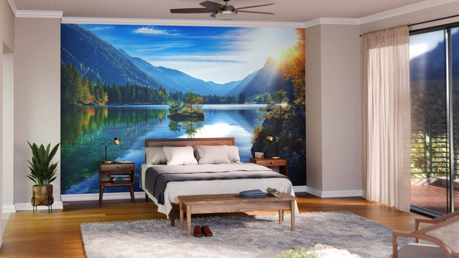 A cozy bedroom with a large bed and wooden furniture features the Hintersee Lake Autumn Sunset Wall Mural. Sunlight filters through sheer curtains, and green plants bring a touch of nature to the modern space.
