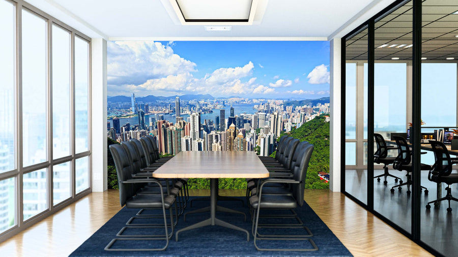 A modern conference room features a long wooden table, black chairs, glass walls, and the Hong Kong View From Peak Wall Mural showing a vibrant city skyline under a blue sky. Sunlight streams in from large windows on the left.