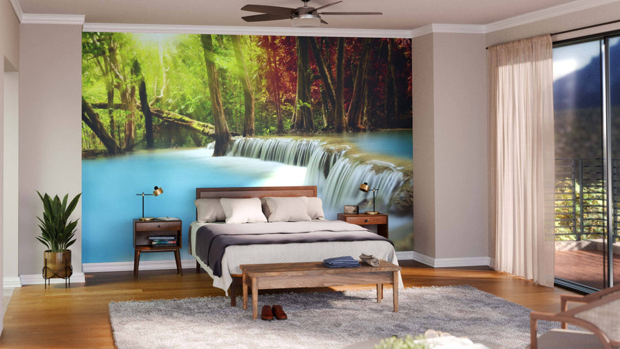 A cozy bedroom features the Huai Mae Kamin Waterfall Wall Mural behind a wooden bed with white and gray bedding. Wooden floors, indoor plants, and large windows with sheer curtains let in plenty of sunlight.