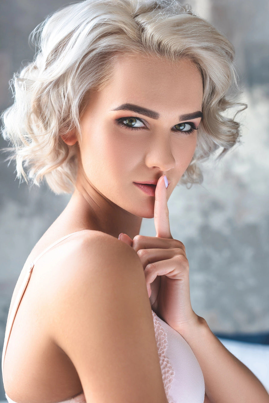 A woman with short, curly blond hair poses thoughtfully with a finger over her lips, suggesting silence. Her subtle eyeliner and natural lip color enhance her quiet sophistication, dressed in light tones against a backdrop reminiscent of the Hush Now Wall Mural.