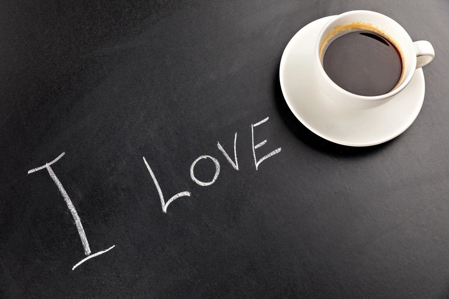 A modern café with wooden tables and stools features the I Love Coffee Wall Mural on a black wall, showing a large coffee cup and “I LOVE.” Small plants decorate each table, while pendant lights add to the cozy atmosphere.