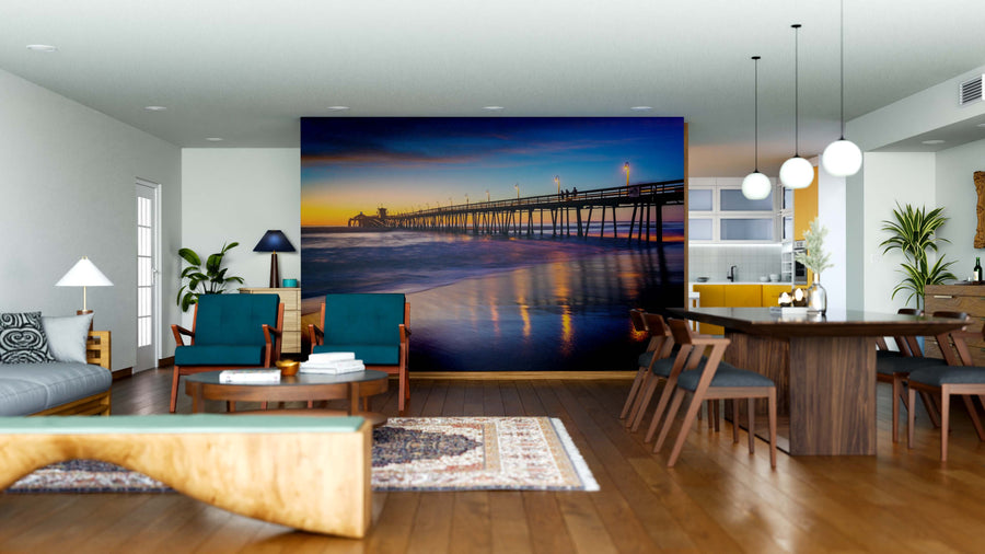 A modern living-dining room with wooden floors and stylish furniture features the Imperial Beach Wall Mural, showcasing a sunset-lit pier over the ocean. Teal chairs, pendant lights, and indoor plants complete the cozy atmosphere.