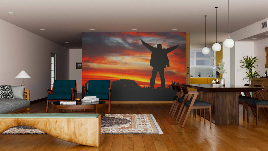 A modern living-dining room features wood floors, blue chairs, pendant lights above the dining table, and the Adoration of Heaven Wall Mural showing a silhouette with raised arms against an orange sunset sky. A small rug sits near the coffee table.