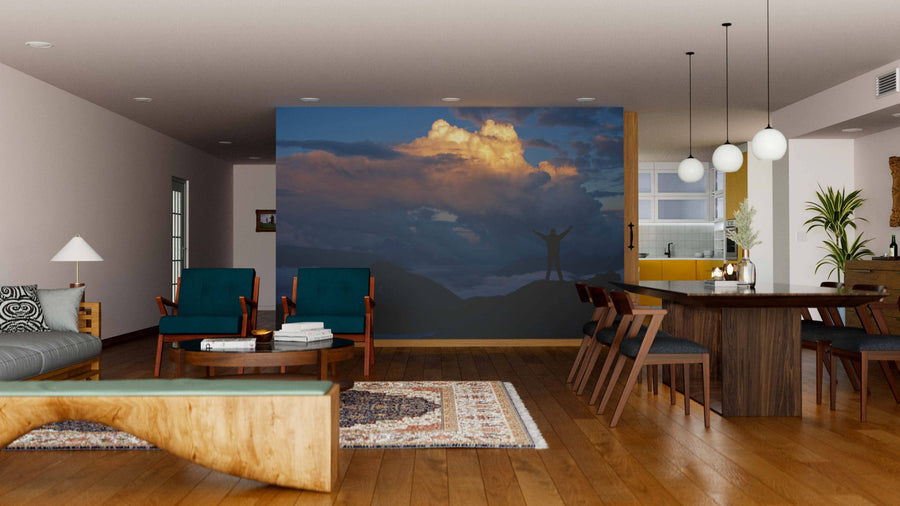 A modern living-dining room features wood floors, mixed seating, and a large Beautiful Cloudscape Wall Mural with a silhouetted figure against a sunset sky. Pendant lights hang above the dining table; the kitchen is visible in the background.