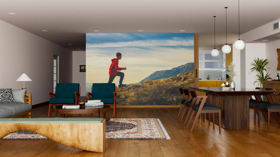 A modern living-dining area with wood floors and furnishings features the "Chasing the Summit Wall Mural," showing a hiker. Blue armchairs, a sofa, a wood dining table, rugs, pendant lights, and an open kitchen create an inviting space.