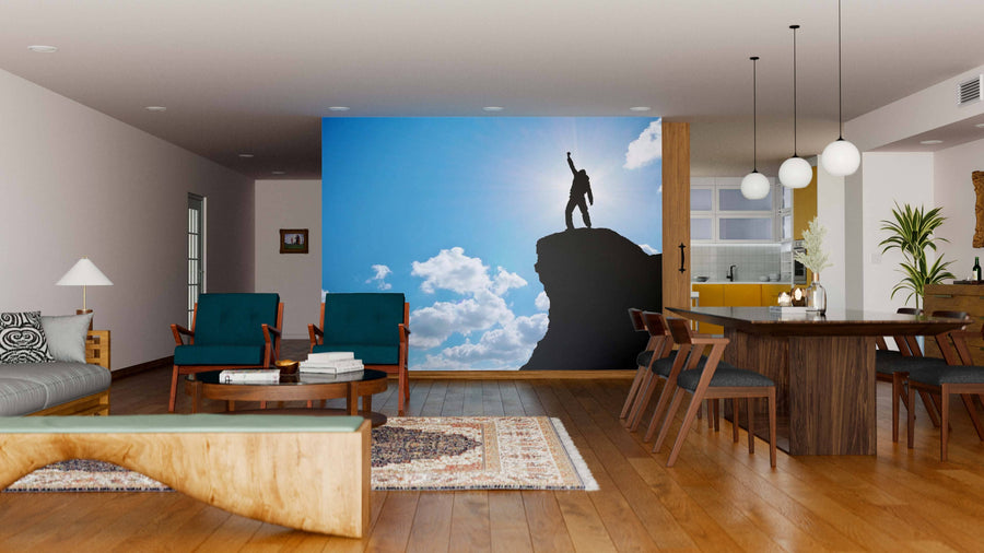 A modern living-dining room with wooden floors, teal chairs, a large wooden dining table, and the Conquering New Heights Wall Mural featuring a silhouette of a person standing triumphantly on a mountain peak against a bright, cloudy sky.
