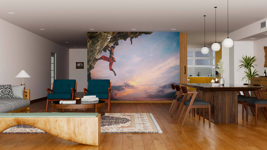 A modern living-dining room with wood floors and furniture features the Defy the Limits Wall Mural of two rock climbers at sunset. Blue armchairs, a large dining table, and pendant lights add to the cozy, eclectic vibe.