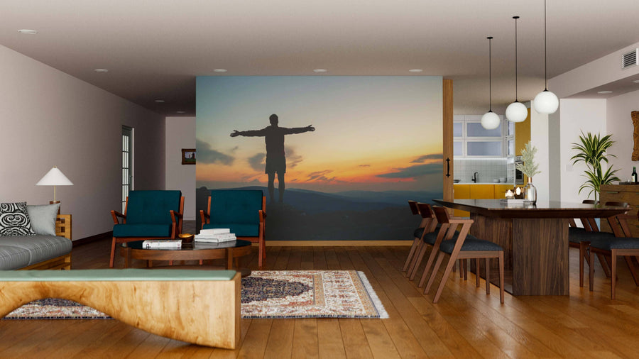 A modern living-dining room with wooden floors, teal chairs, and globe pendant lights is enhanced by the Embracing the Sunset Wall Mural, which adds warmth and depth with its silhouette against a sunset landscape.