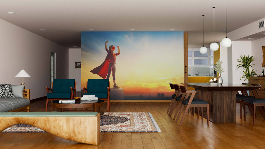 A modern open-plan living/dining room with wood floors, teal armchairs, and the "Fearless and Free Wall Mural" featuring a child in a red cape posing as a superhero against a sunset sky for playful, contemporary style.