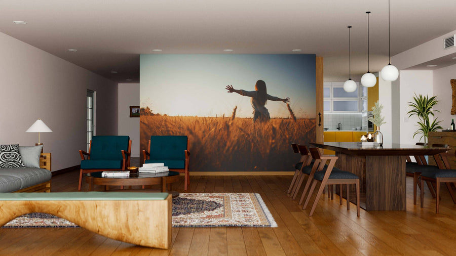 Modern living-dining room with wooden floors and furniture. A Field of Light Wall Mural depicting a person with outstretched arms at sunset is the focal point, complemented by teal chairs, wood dining table, gray sofa, rugs, and globe lights.