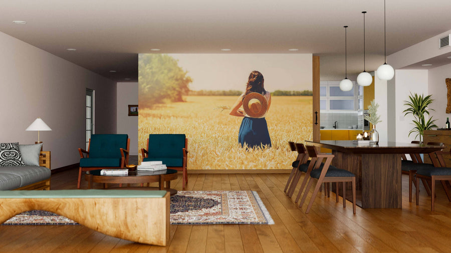 A modern living-dining room features wooden floors, a gray sofa, green armchairs, and a Fields of Solitude Wall Mural depicting a woman in a hat among wheat. Pendant lights hang over the dining table; the kitchen is visible in the background.