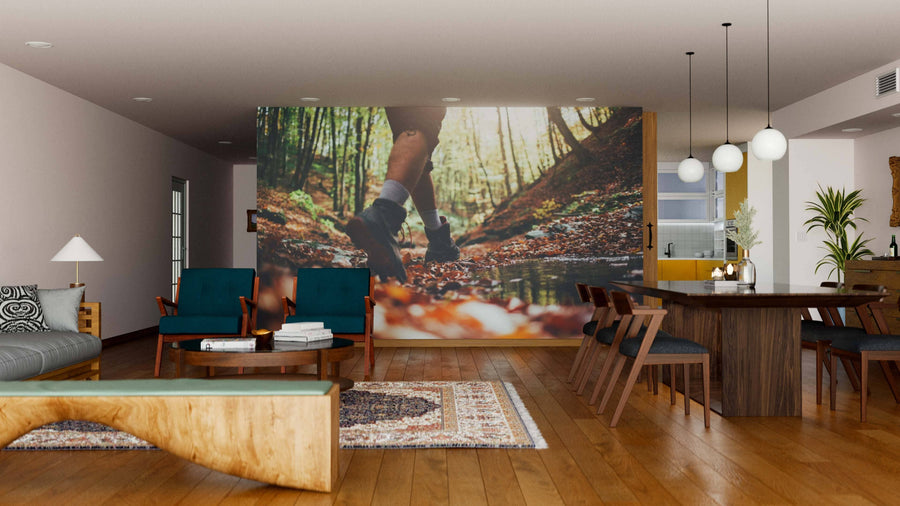 A modern living and dining area with wooden floors, stylish furniture, teal chairs, pendant lights, an open kitchen in the background, and a large Footsteps Forward Wall Mural featuring a person hiking in a forest.