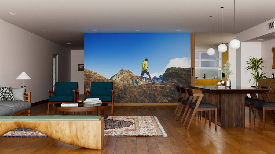 A modern living-dining room features wooden floors, stylish furniture, and the "Freedom in Motion Wall Mural" depicting a person hiking in mountains under a blue sky. Warm lighting and contemporary decor complete the cozy, open ambiance.
